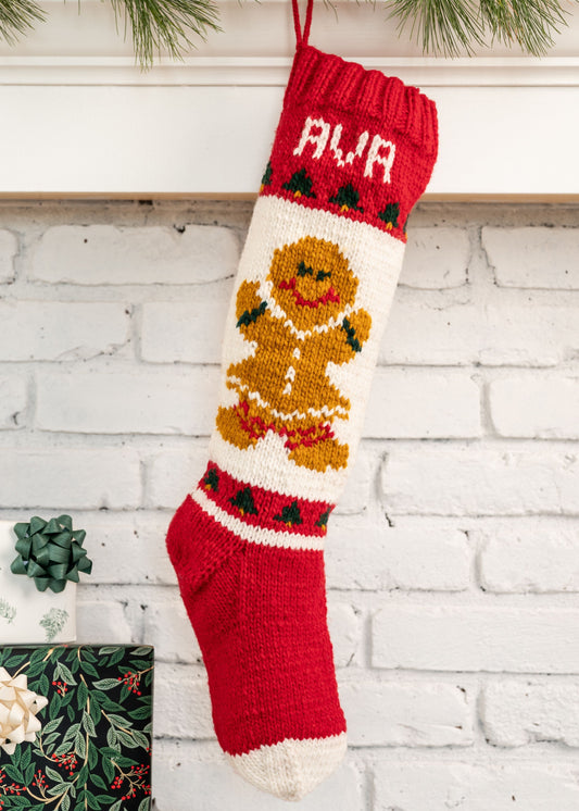 Knitted Christmas stocking with gingerbread girl design, hanging against a white brick wall.